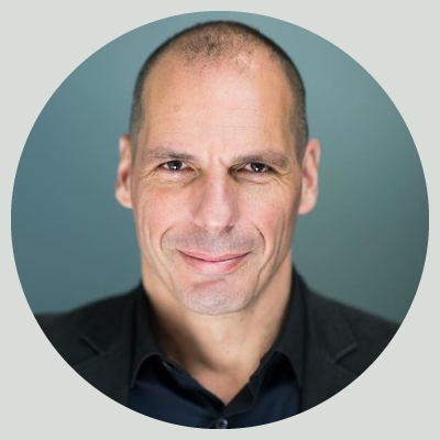 Portrait of Yanis Varoufakis. He is wearing a dark blue shirt and a black jacket. He is staring optimistically and smiling