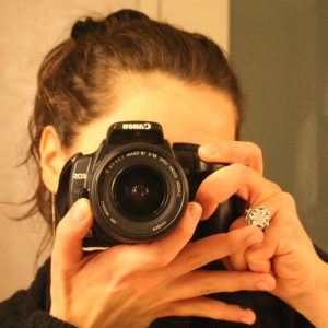 Artistic portrait of Chara Ioannou. She has brown hair in a ponytail and is wearing a black pullover. Her face is hiding behind her Canon camera which she is holding with both hands. She is wearing a bright ring.