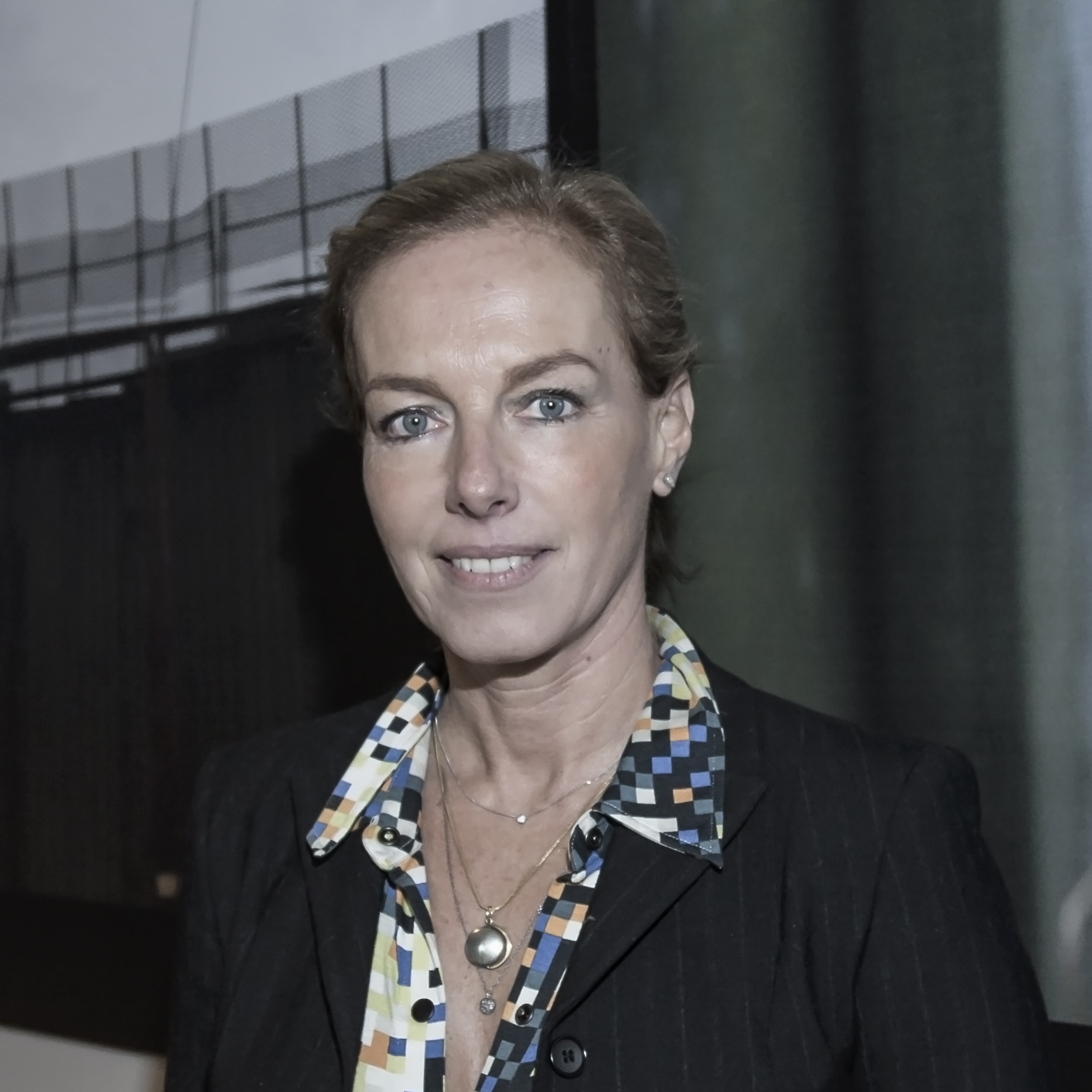 Portrait of Danae Stratou She has blond hair in a ponytail and is wearing a colorful patterned shirt and a black jacket She is staring and smiling broadly