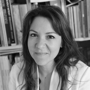 Black and white portrait of Iliana Fokianaki. She has long black hair and brown eyes. She is wearing a white shirt and is smiling at the camera, posing in a bookcase background.