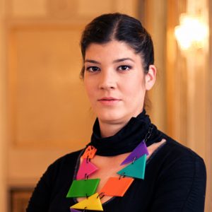 Portrait of Ivana Nenadović She has dark hair in a ponytail and is wearing a black blouse and a sophisticated necklace with big colorful triangles She is staring