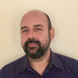 Portrait of Nick Kanarelis. He has dark hair moustache and beard. He is wearing a blue shirt. He is staring and smiling.