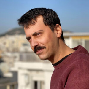 Portrait of George Tsalikis in a blurred city background and a hill. He has short black hair and a moustache. He is wearing a burgundy blouse. He is gazing in the distance.